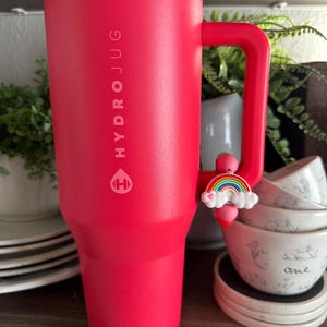 May include: A large, red Hydrojug tumbler with a handle. The tumbler has the Hydrojug logo and text printed vertically. A rainbow charm hangs from the handle. The tumbler is on a shelf with other items.