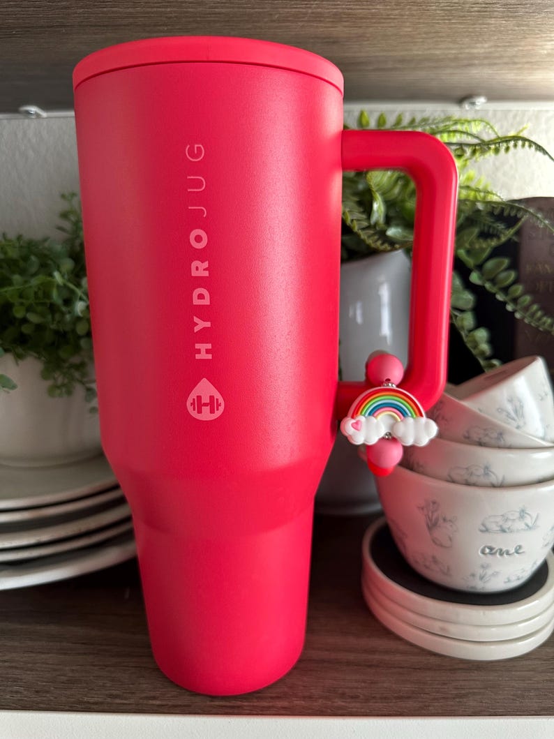 May include: A large, red Hydrojug tumbler with a handle. The tumbler has the Hydrojug logo and text printed vertically. A rainbow charm hangs from the handle. The tumbler is on a shelf with other items.