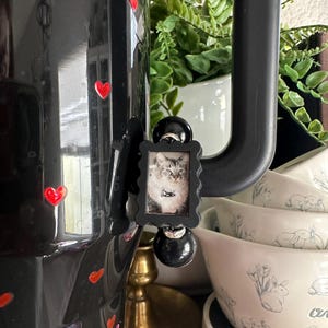 May include: A black decorative coffee pot with red heart accents and a small framed picture of a cat. The pot is near a stack of white bowls with a bunny and floral design.