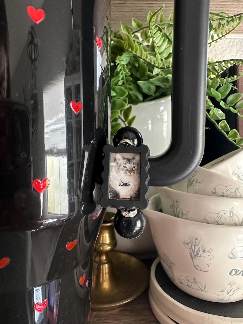 May include: A black decorative coffee pot with red heart accents and a small framed picture of a cat. The pot is near a stack of white bowls with a bunny and floral design.