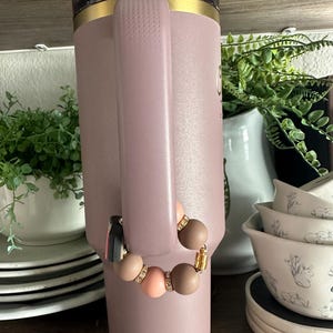 Puede incluir: Un vaso aislante grande de color p&uacute;rpura claro con asa, ribete dorado y tapa transparente. Una pulsera de cuentas con cuentas marrones, melocot&oacute;n y beige est&aacute; sujeta al asa. La pulsera tiene detalles dorados y una borla negra.