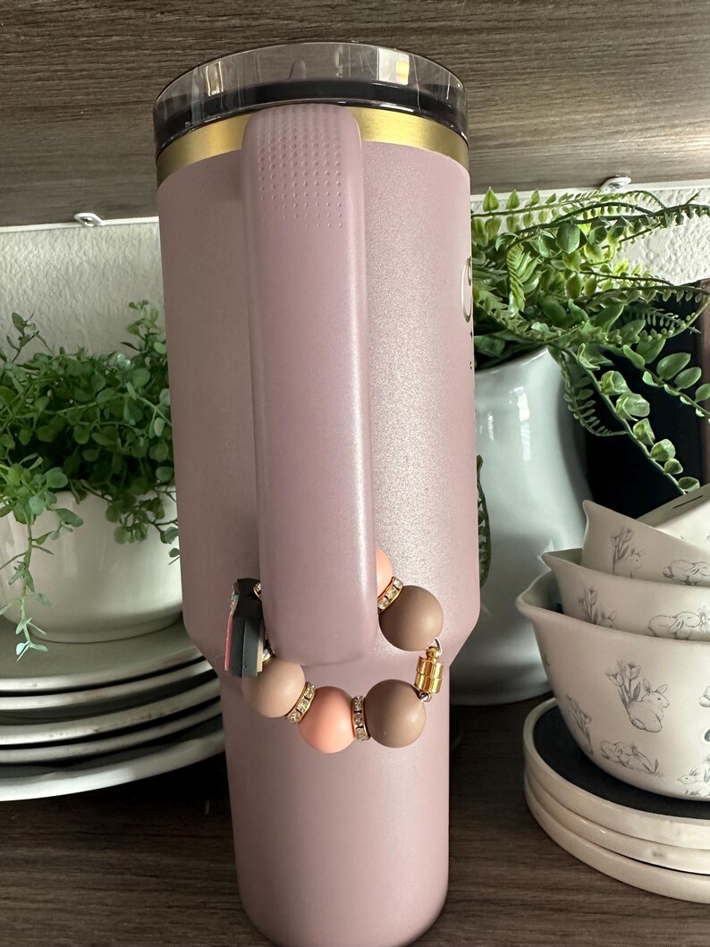 Puede incluir: Un vaso aislante grande de color p&uacute;rpura claro con asa, ribete dorado y tapa transparente. Una pulsera de cuentas con cuentas marrones, melocot&oacute;n y beige est&aacute; sujeta al asa. La pulsera tiene detalles dorados y una borla negra.