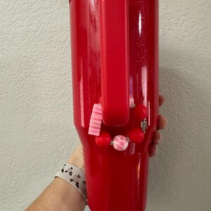 Puede incluir: Un vaso rojo cubierto de purpurina con asa. El vaso est&aacute; adornado con una pulsera de cuentas rosas y rojas. El vaso lo sostiene una persona.