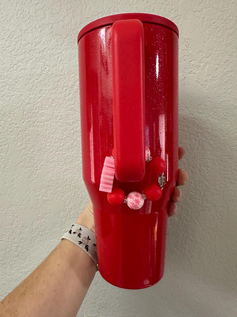 Puede incluir: Un vaso rojo cubierto de purpurina con asa. El vaso est&aacute; adornado con una pulsera de cuentas rosas y rojas. El vaso lo sostiene una persona.