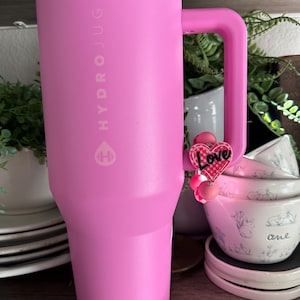 May include: A large, pink Hydrojug water bottle with a handle. The bottle has the Hydrojug logo and text on the side. A pink charm with the word "Love" hangs from the handle. The background includes white bowls and greenery.