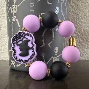 May include: A Halloween-themed bracelet featuring lavender and black beads. The bracelet includes a charm with a witch silhouette and spiderweb design. Gold-colored accents connect the beads, creating a festive accessory.