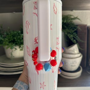 May include: White insulated tumbler with a handle, adorned with red seashell and seahorse illustrations. A red, blue, and silver beaded bracelet is attached to the handle. The tumbler has a white lid.