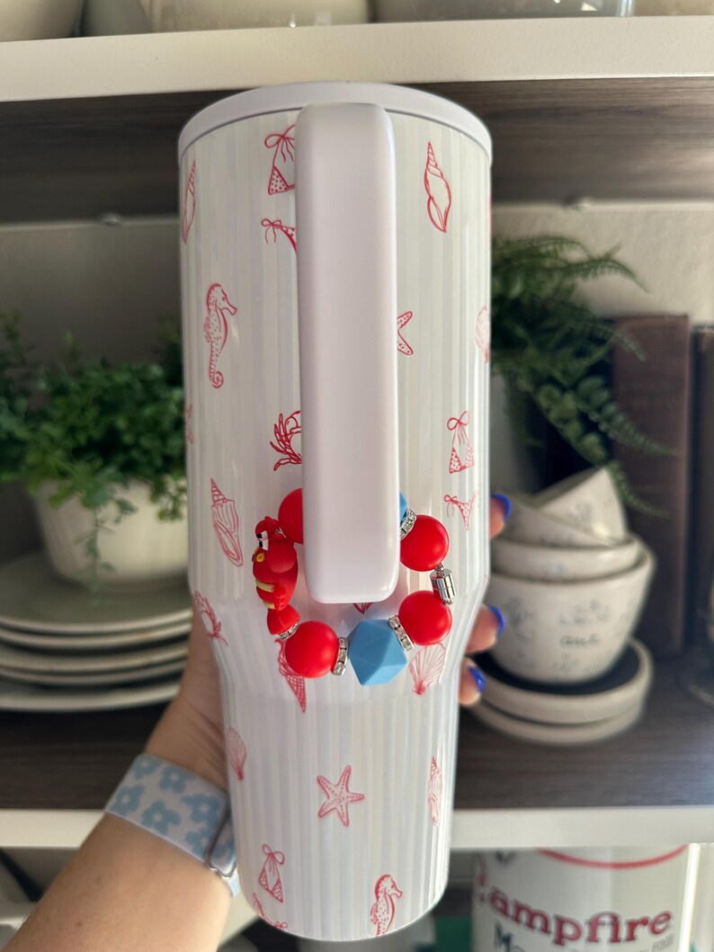 May include: White insulated tumbler with a handle, adorned with red seashell and seahorse illustrations. A red, blue, and silver beaded bracelet is attached to the handle. The tumbler has a white lid.