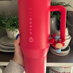 May include: A large, red HydroJug water bottle with a handle and a crab charm. The bottle has the HydroJug logo and text printed on it. The handle has a crab charm attached to it.