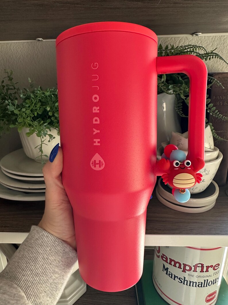May include: A large, red HydroJug water bottle with a handle and a crab charm. The bottle has the HydroJug logo and text printed on it. The handle has a crab charm attached to it.