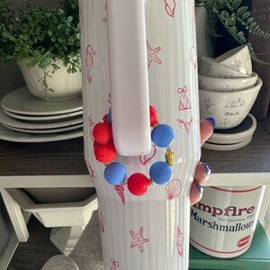 Puede incluir: Vaso blanco con asa, decorado con ilustraciones rojas de conchas marinas y estrellas de mar. Una pulsera de cuentas rojas, blancas y azules est&aacute; sujeta al asa. El vaso es cil&iacute;ndrico y aislado.