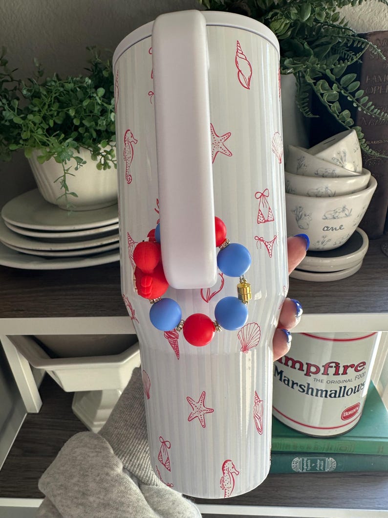 Puede incluir: Vaso blanco con asa, decorado con ilustraciones rojas de conchas marinas y estrellas de mar. Una pulsera de cuentas rojas, blancas y azules est&aacute; sujeta al asa. El vaso es cil&iacute;ndrico y aislado.