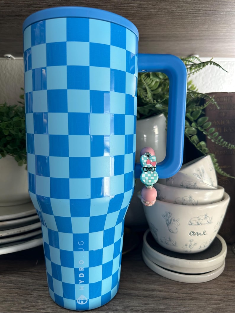 Op de afbeelding: Een blauw en lichtblauw geblokte Hydro Jug-beker met een blauw handvat. Een decoratieve bedel met een konijnenontwerp is aan het handvat bevestigd. De beker is ontworpen om dranken koud te houden.