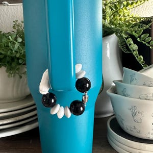 May include: A turquoise tumbler with a black and white beaded bracelet attached to the handle. The bracelet features black and white beads. Other items in the background include white dishes and greenery.