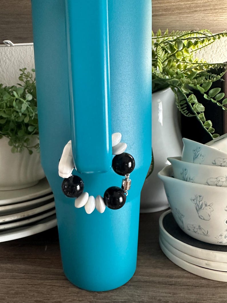 May include: A turquoise tumbler with a black and white beaded bracelet attached to the handle. The bracelet features black and white beads. Other items in the background include white dishes and greenery.