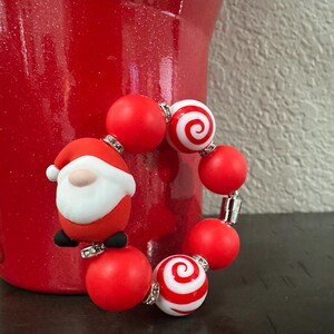 Może przedstawiać: Świąteczna bransoletka z czerwonymi i białymi koralikami, w tym dwoma ze spiralnym wzorem. W zestawie znajduje się r&oacute;wnież mała figurka Świętego Mikołaja. Bransoletka znajduje się obok czerwonego wazonu. To świąteczny dodatek.