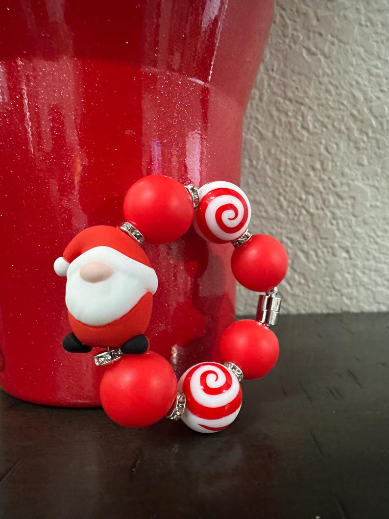 Może przedstawiać: Świąteczna bransoletka z czerwonymi i białymi koralikami, w tym dwoma ze spiralnym wzorem. W zestawie znajduje się r&oacute;wnież mała figurka Świętego Mikołaja. Bransoletka znajduje się obok czerwonego wazonu. To świąteczny dodatek.