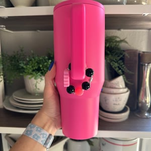 Pode incluir: Um copo t&eacute;rmico rosa brilhante com uma al&ccedil;a. Um berloque com contas pretas e rosas est&aacute; pendurado na al&ccedil;a. O copo &eacute; exibido em uma prateleira com outros utens&iacute;lios de cozinha e itens decorativos.