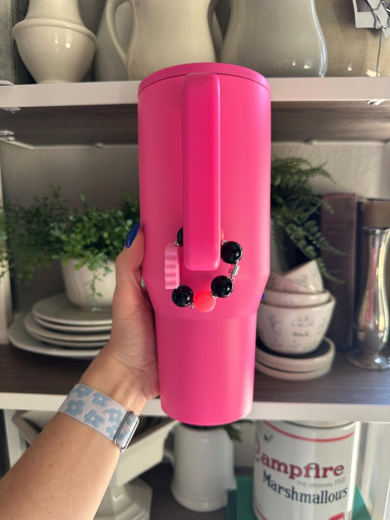 Pode incluir: Um copo t&eacute;rmico rosa brilhante com uma al&ccedil;a. Um berloque com contas pretas e rosas est&aacute; pendurado na al&ccedil;a. O copo &eacute; exibido em uma prateleira com outros utens&iacute;lios de cozinha e itens decorativos.