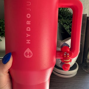May include: A large, red HydroJug water bottle with a matching handle. The bottle features the HydroJug logo and a small, red lobster charm attached to the handle. The bottle is held by a person with blue nail polish.