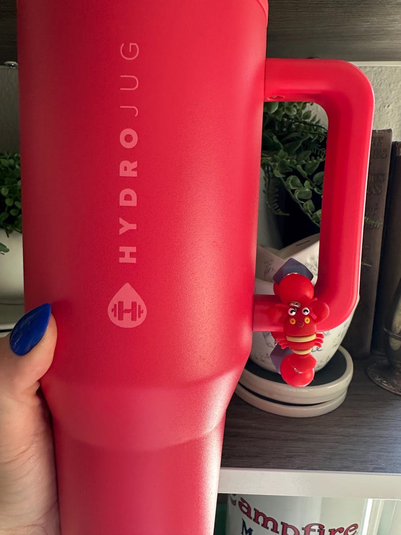 May include: A large, red HydroJug water bottle with a matching handle. The bottle features the HydroJug logo and a small, red lobster charm attached to the handle. The bottle is held by a person with blue nail polish.