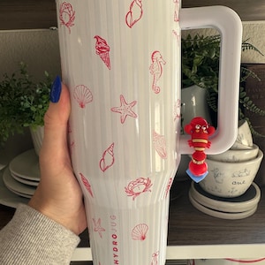 以下が含まれることがあります： 大きなハンドルが付いた白いHydroJug。赤い貝殻、ヒトデ、アイスクリームコーンのイラストで装飾されています。ジャグには垂直の溝があり、側面には「HYDROJUG」の文字が印刷されています。赤いロブスターチャームがハンドルからぶら下がっています。