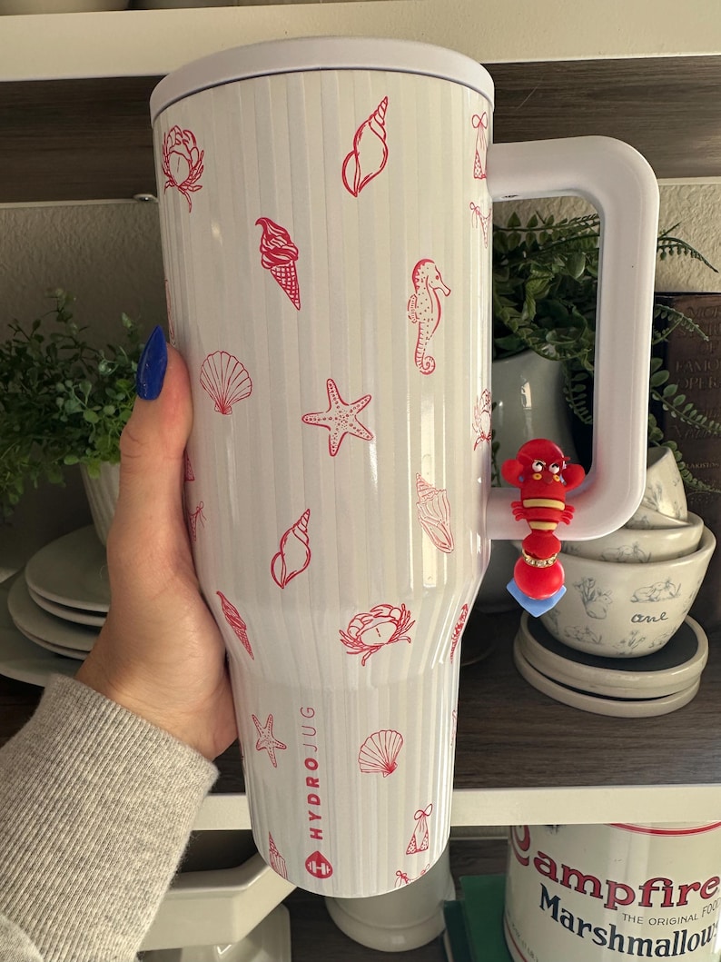 以下が含まれることがあります： 大きなハンドルが付いた白いHydroJug。赤い貝殻、ヒトデ、アイスクリームコーンのイラストで装飾されています。ジャグには垂直の溝があり、側面には「HYDROJUG」の文字が印刷されています。赤いロブスターチャームがハンドルからぶら下がっています。