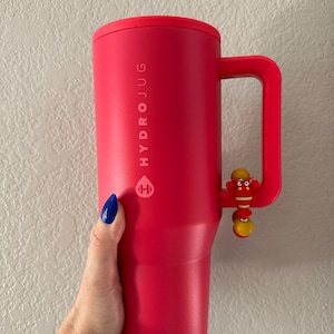 May include: A vibrant red HydroJug water bottle with a handle and a decorative charm. The bottle features the HydroJug logo and text. The handle has a small, colorful charm attached. The bottle is held by a hand with blue nail polish.