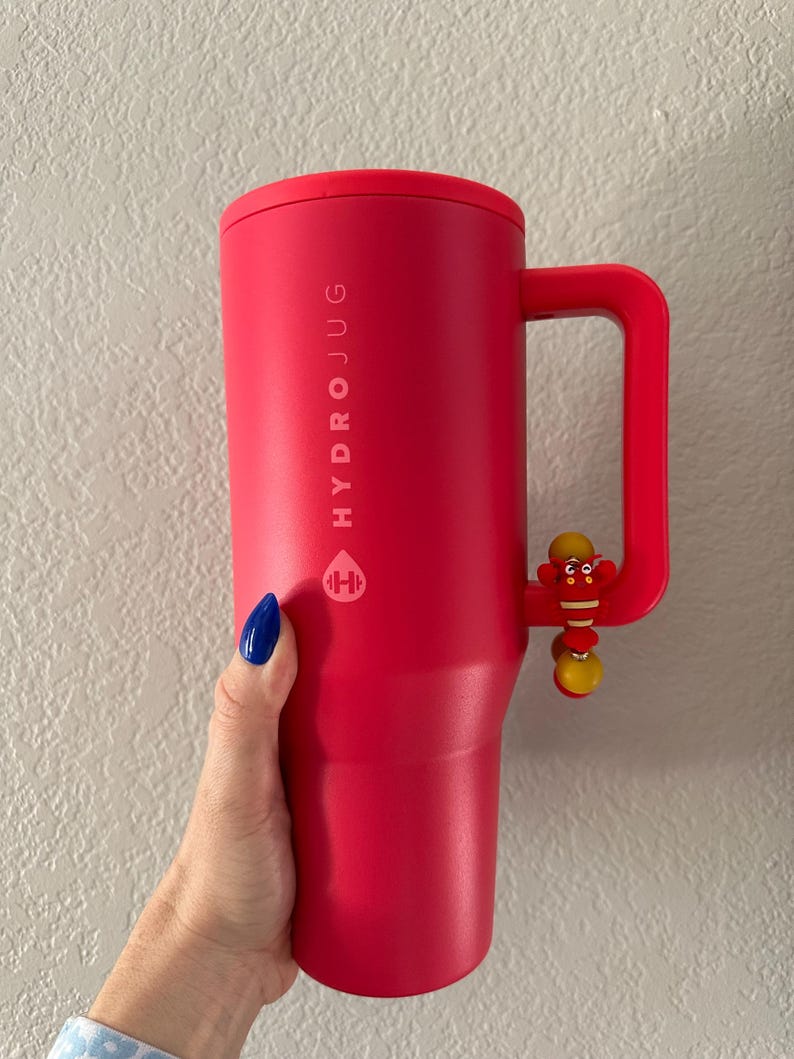 May include: A vibrant red HydroJug water bottle with a handle and a decorative charm. The bottle features the HydroJug logo and text. The handle has a small, colorful charm attached. The bottle is held by a hand with blue nail polish.