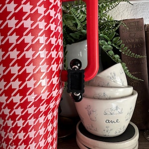 May include: A red and white houndstooth patterned tumbler with a red handle. Stacked white bowls with blue bunny illustrations and the word "one" are visible. The items are on a wooden shelf.