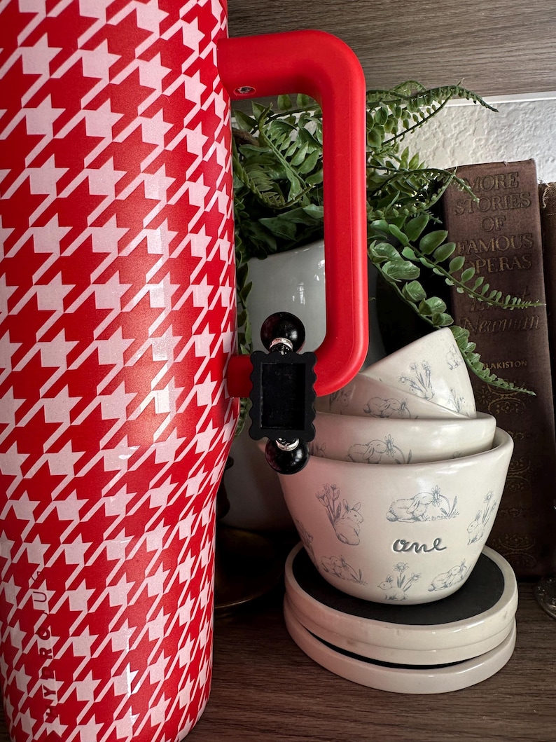 May include: A red and white houndstooth patterned tumbler with a red handle. Stacked white bowls with blue bunny illustrations and the word "one" are visible. The items are on a wooden shelf.