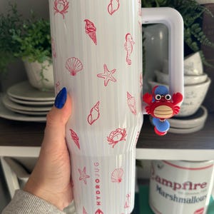 Puede incluir: HydroJug blanco con dise&ntilde;os rojos de conchas marinas, conos de helado y estrellas de mar. La jarra tiene un asa blanca y un dije de cangrejo rojo. El texto "HYDROJUG" est&aacute; impreso en el lateral.