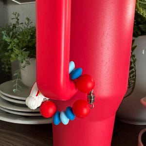 May include: A vibrant red tumbler with a magnetic charm bracelet attached. The bracelet features red and blue beads, and a silver magnetic clasp. The tumbler is cylindrical with a handle.