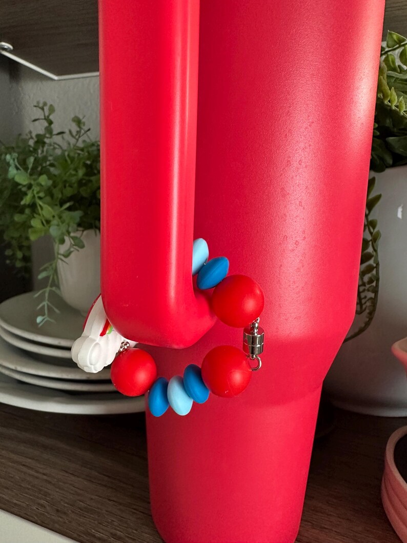May include: A vibrant red tumbler with a magnetic charm bracelet attached. The bracelet features red and blue beads, and a silver magnetic clasp. The tumbler is cylindrical with a handle.