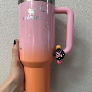 May include: A pink and orange Stanley tumbler with a handle. The tumbler has a gradient design, with a pink top that fades into orange. It features a silver logo and small holographic accents. A pin that says "Spook Babe" is attached to the handle.