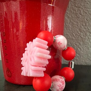 Puede incluir: Una pulsera festiva con cuentas rojas y rosas. La pulsera incluye un dije en forma de copo de nieve rosa claro y cuentas rojas y rosas alternas con un patr&oacute;n de copo de nieve. El fondo es rojo.