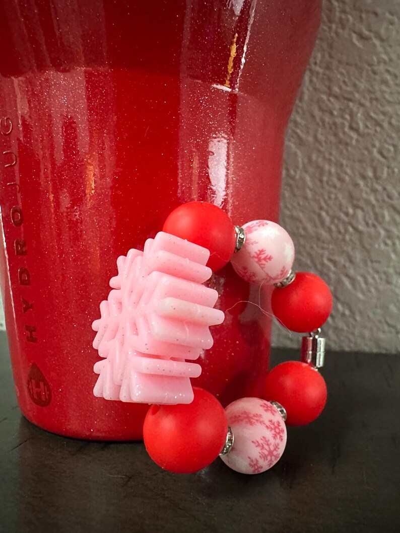 Puede incluir: Una pulsera festiva con cuentas rojas y rosas. La pulsera incluye un dije en forma de copo de nieve rosa claro y cuentas rojas y rosas alternas con un patr&oacute;n de copo de nieve. El fondo es rojo.