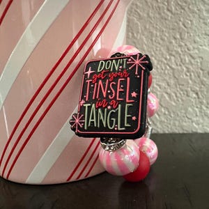 May include: A Christmas-themed charm bracelet with pink and red beads. The focal point is a black rectangular charm with the text "DON'T get your TINSEL in a TANGLE" in pink and green.