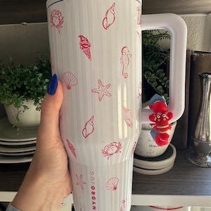 May include: White HydroJug with a white handle and lid, decorated with red seashell, starfish, and ice cream cone illustrations. The HydroJug has the brand name printed on the side. A red lobster charm is attached to the handle.