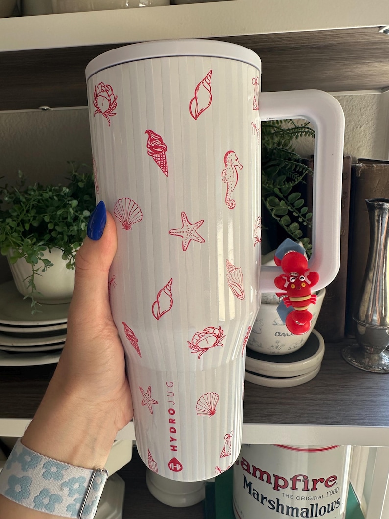 May include: White HydroJug with a white handle and lid, decorated with red seashell, starfish, and ice cream cone illustrations. The HydroJug has the brand name printed on the side. A red lobster charm is attached to the handle.