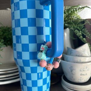 Op de afbeelding: Een blauw en lichtblauw geruite beker met een blauwe handgreep. Een kralenbedel met roze en patronen kralen en een lichtblauw konijn met een bril hangt aan de handgreep. De beker staat op een houten oppervlak.