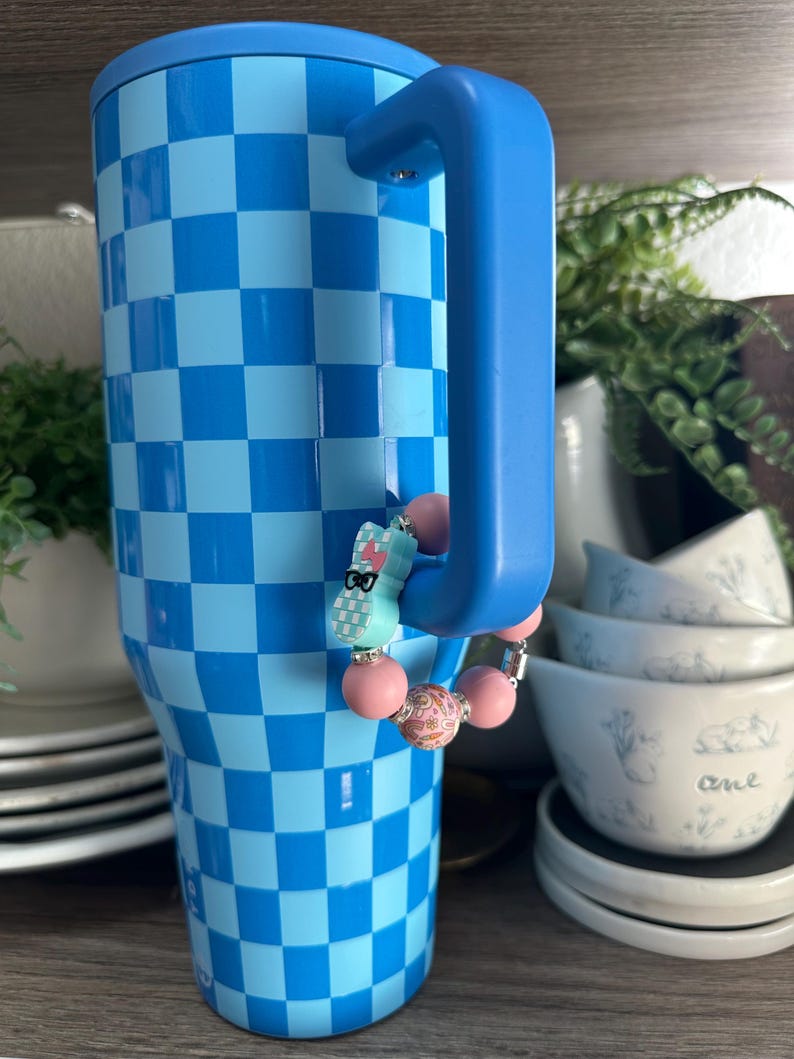 Op de afbeelding: Een blauw en lichtblauw geruite beker met een blauwe handgreep. Een kralenbedel met roze en patronen kralen en een lichtblauw konijn met een bril hangt aan de handgreep. De beker staat op een houten oppervlak.