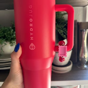 May include: A red Hydrojug water bottle with a handle. The bottle has the word "HYDROJUG" printed vertically on the side, along with a logo. A pink charm is attached to the handle. The bottle is cylindrical and appears to be made of insulated material.