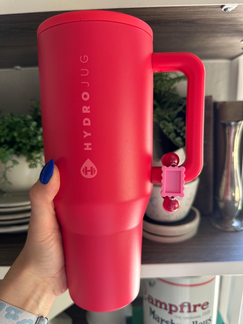 May include: A red Hydrojug water bottle with a handle. The bottle has the word "HYDROJUG" printed vertically on the side, along with a logo. A pink charm is attached to the handle. The bottle is cylindrical and appears to be made of insulated material.