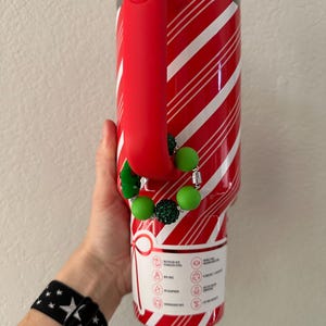 Pode incluir: Um copo de a&ccedil;o inoxid&aacute;vel listrado em vermelho e branco com uma al&ccedil;a vermelha. O copo tem um berloque de contas verdes e prateadas. Uma pulseira preta com estrelas brancas &eacute; vis&iacute;vel.