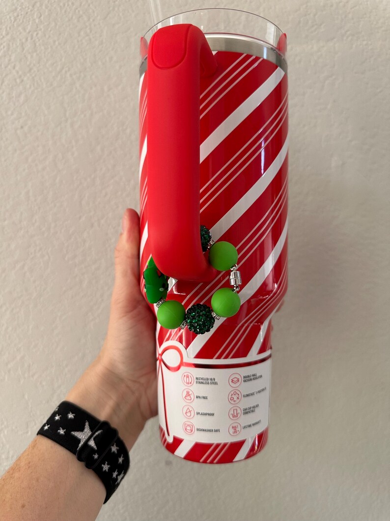 Pode incluir: Um copo de a&ccedil;o inoxid&aacute;vel listrado em vermelho e branco com uma al&ccedil;a vermelha. O copo tem um berloque de contas verdes e prateadas. Uma pulseira preta com estrelas brancas &eacute; vis&iacute;vel.