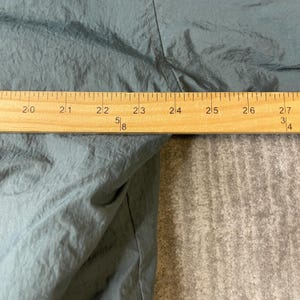 May include: A dark gray, crinkled fabric garment is positioned behind a wooden ruler. The ruler shows a measurement spanning from 20 inches to 27 inches, clearly indicating the width of the fabric section visible in this close-up view.