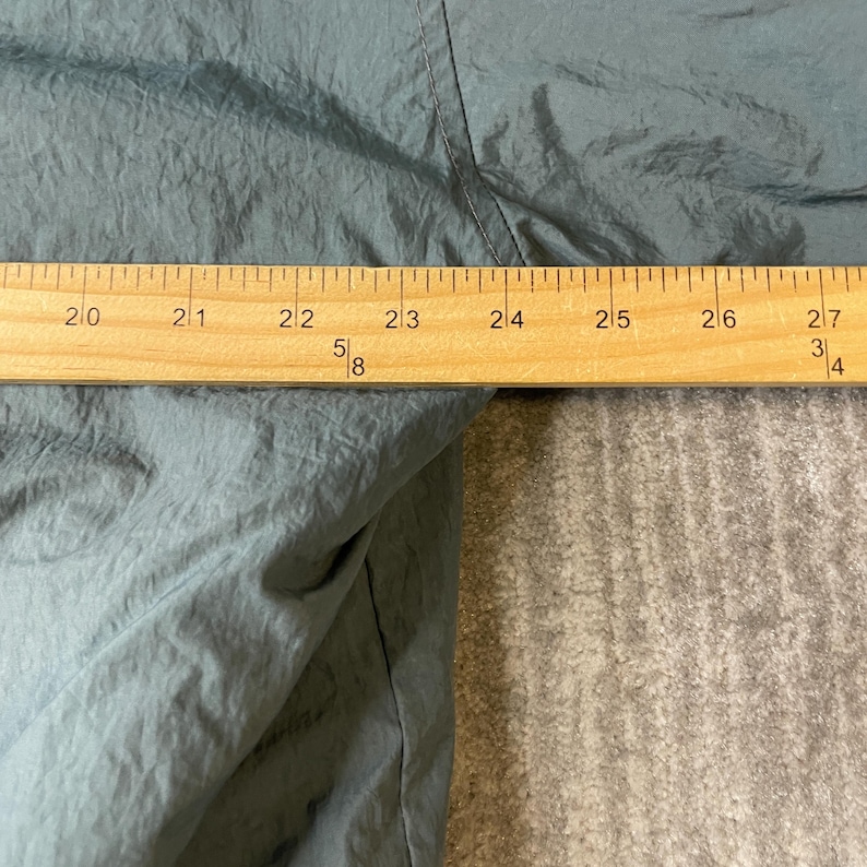May include: A dark gray, crinkled fabric garment is positioned behind a wooden ruler. The ruler shows a measurement spanning from 20 inches to 27 inches, clearly indicating the width of the fabric section visible in this close-up view.