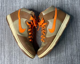 May include: Pair of high-top trainers in brown, tan, and orange. The shoes feature a tan base with brown overlays and a large orange swoosh on the side. Orange laces complete the design. The soles are off-white.