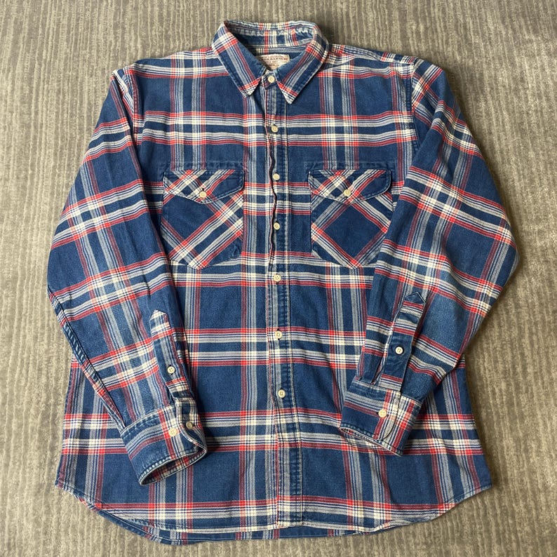 May include: A long-sleeved, button-down shirt in a classic plaid pattern. The shirt features a blue base with red and white intersecting lines, creating a checkered design. It has two chest pockets, a button-down collar, and buttoned cuffs. The shirt is made of a soft, woven fabric.